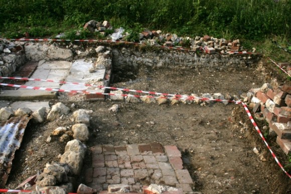 View N from front door across paved path, with possible earth floored yard to its right, toilet top left and garden top right