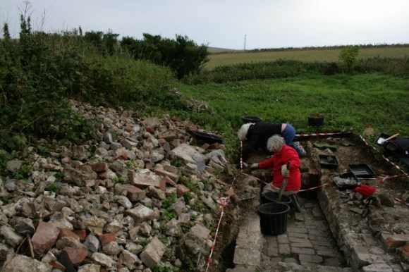 Looking S at an excavation of the east side of a concrete platform beyond the 'woodstore'
