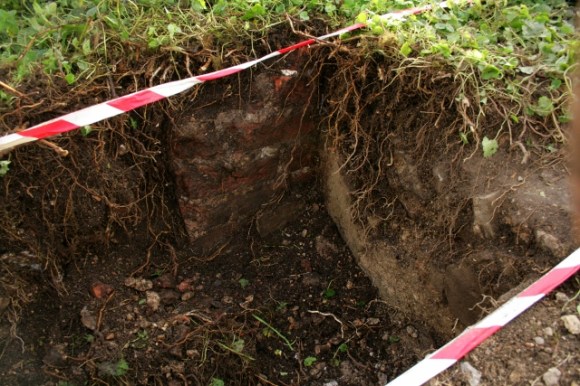 Looking SW from concrete platform at plastered east cottage wall and projecting base of chimney 