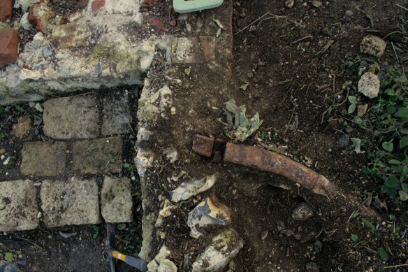 Looking S over water pipe, garden/farmyard wall, stone blocks, and NW corner of cottage walls