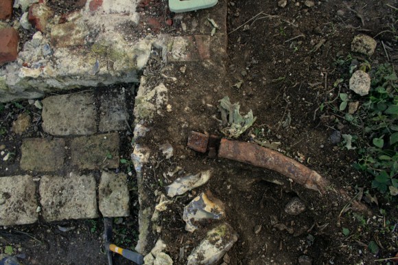 Looking S over water pipe, garden/farmyard wall, stone blocks, and NW corner of cottage walls