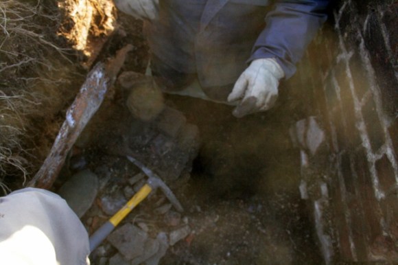 Looking S at trench next to base of fireplace, pulling up edge of sheet of metal embedded in side of trench to see underneath
